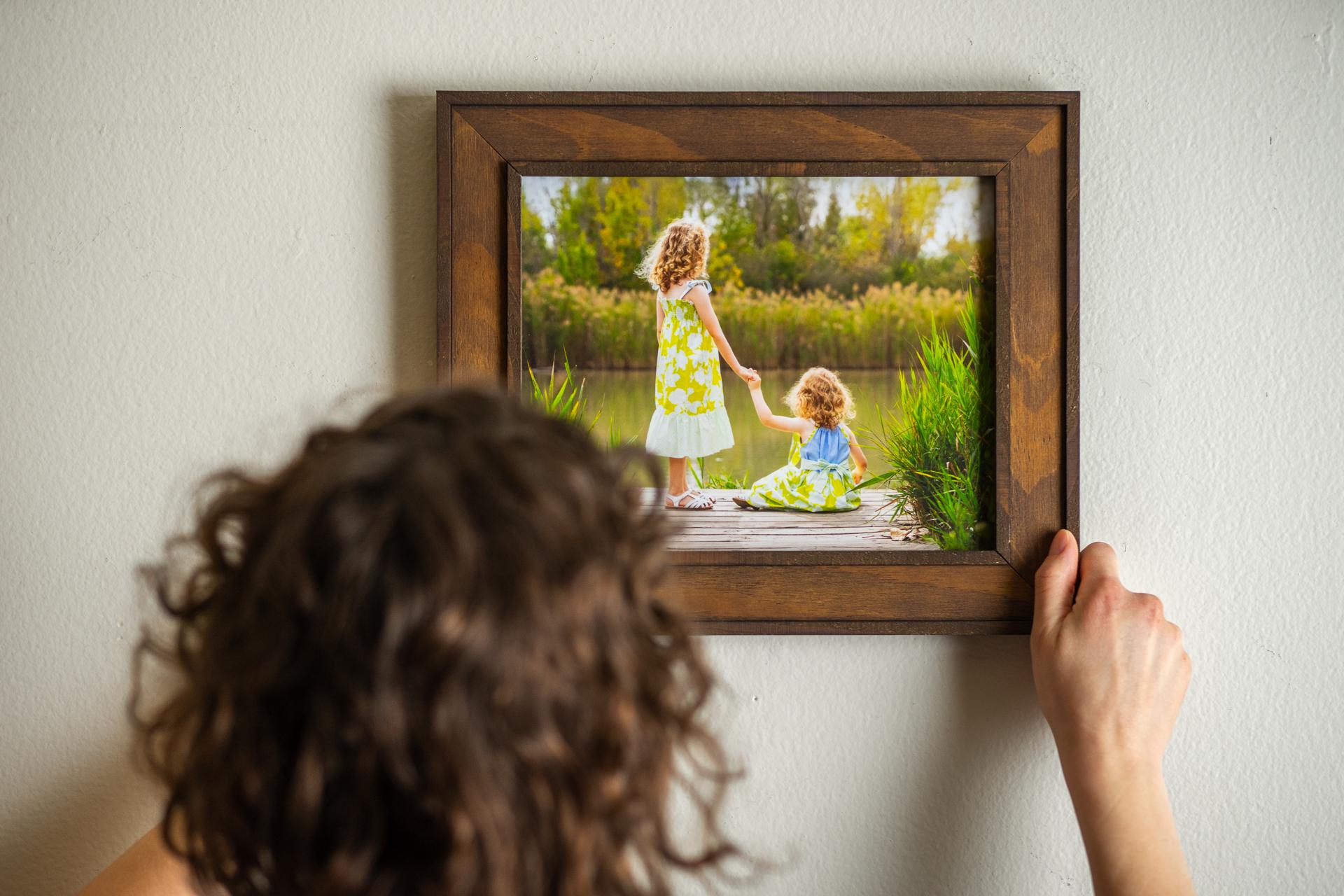 A person hangs a wooden-framed photo of two young girls by a lake on a white wall.