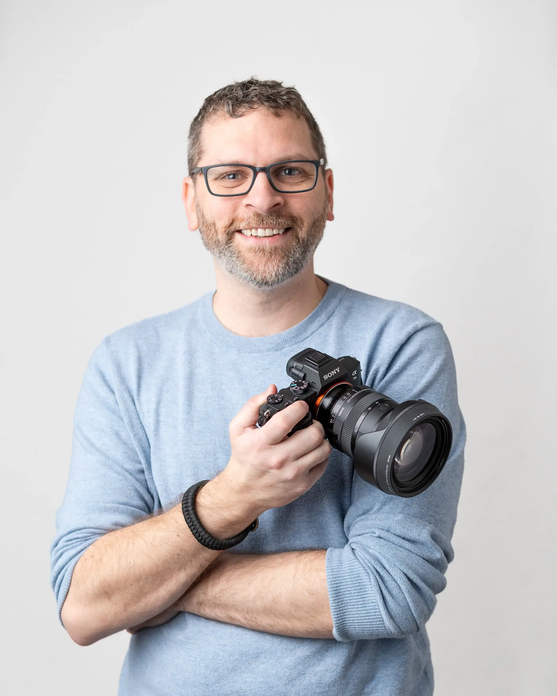 Man with glasses smiling and holding a camera while wearing a light blue sweater against a plain background.