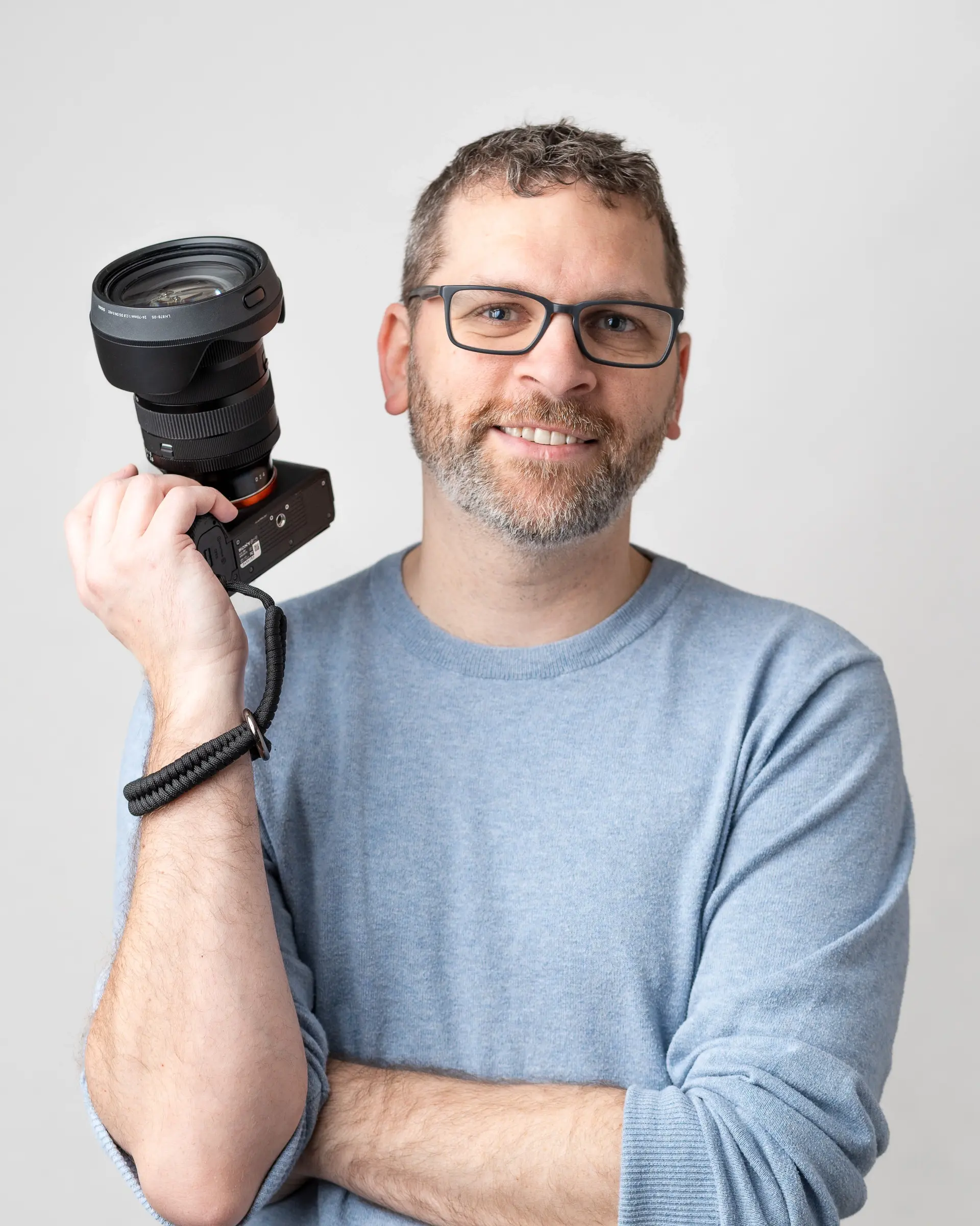 A man with glasses and a beard holds a camera, smiling. He is wearing a light blue sweater against a plain white background.