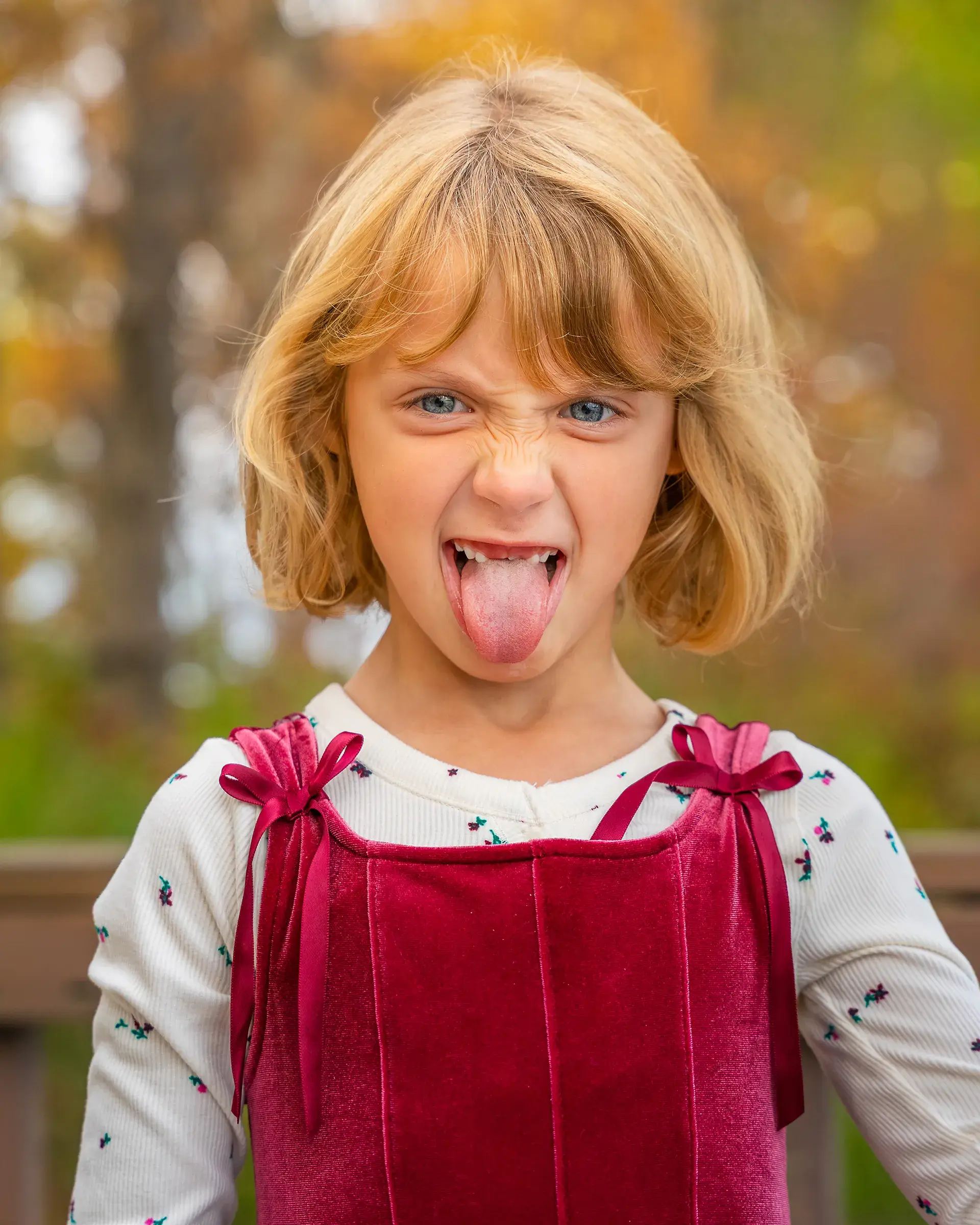 Child with short blonde hair playfully sticks out tongue, wearing a red velvet dress and patterned shirt, with autumn foliage in the background.